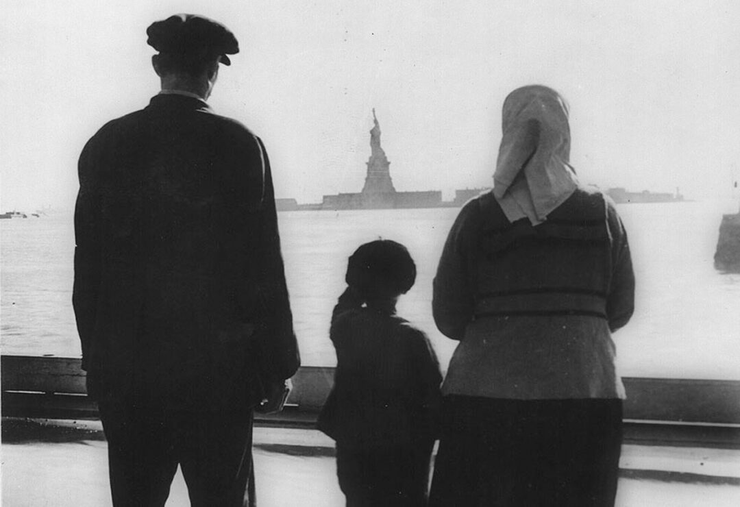 Three people—an adult man, a child, and a woman in a headscarf—stand on a boat looking across the water at the distant Statue of Liberty. The scene suggests immigration or arrival in America.