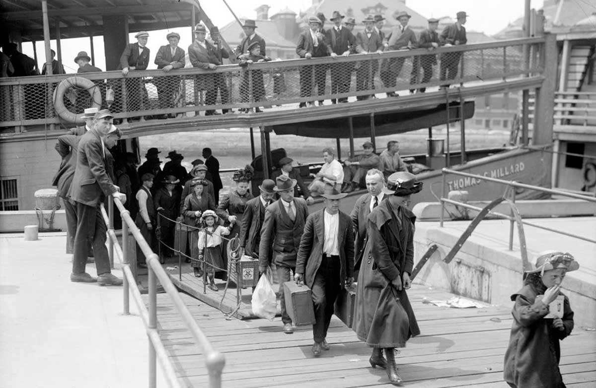 A group of people, including men, women, and children, walk down a gangway from a docked boat, carrying luggage, while others stand and watch from an upper deck. The scene appears to be from the early 20th century.