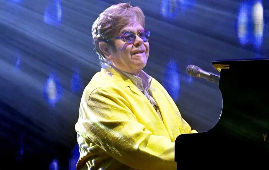 A musician in a yellow jacket and blue glasses plays a black piano on stage, illuminated by blue and white stage lights.