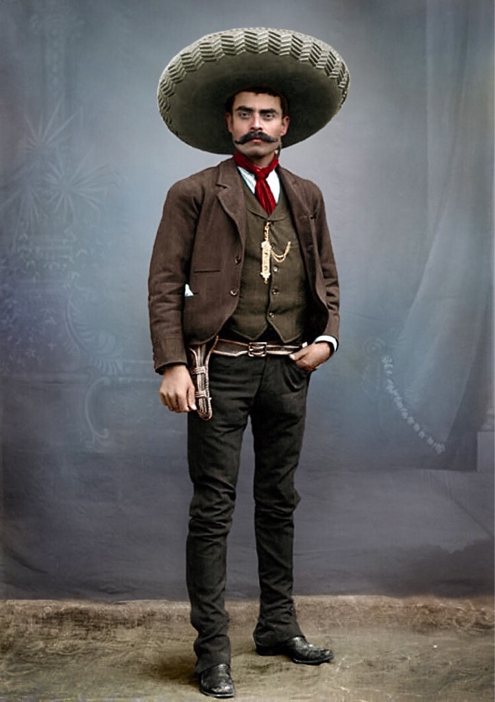 A man wearing a large sombrero, dark suit, vest, and red tie stands confidently with one hand on his hip and a gun holstered at his side, against a vintage studio backdrop.