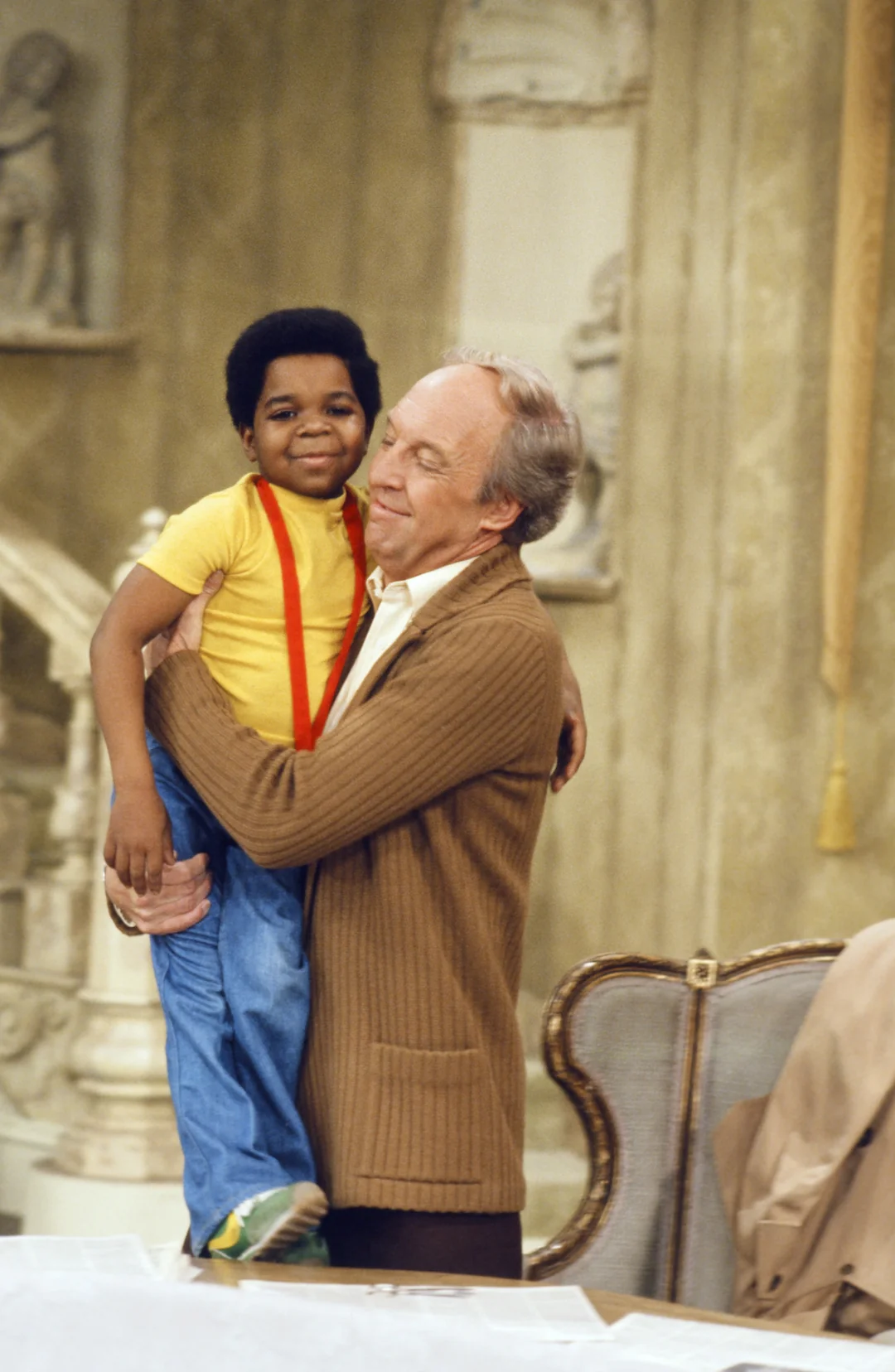 A smiling older man in a brown sweater hugs a young boy wearing a yellow shirt with red suspenders and blue jeans inside a warmly lit, elegant room with ornate decor.