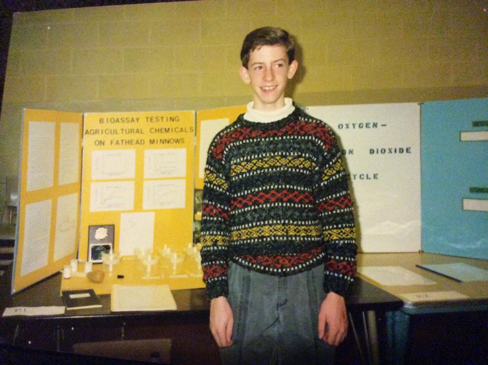 A smiling boy in a patterned sweater stands in front of a science fair project display titled "Bioassay Testing Agricultural Chemicals on Fathead Minnows," featuring charts, papers, and small containers on a table.