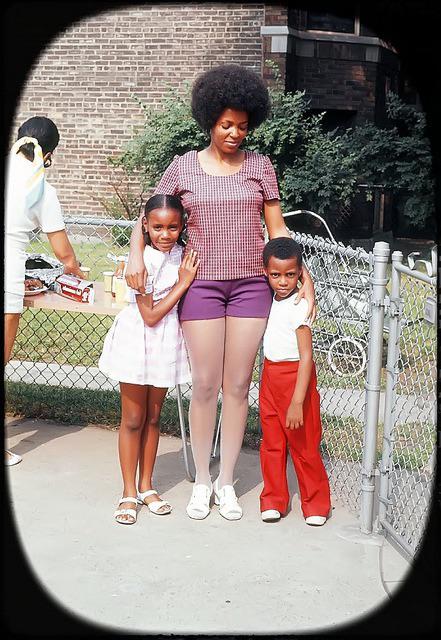 A woman stands outdoors with her arms around a young girl in a pink dress and a young boy in red pants. They are smiling near a chain-link fence with a brick building and trees in the background.