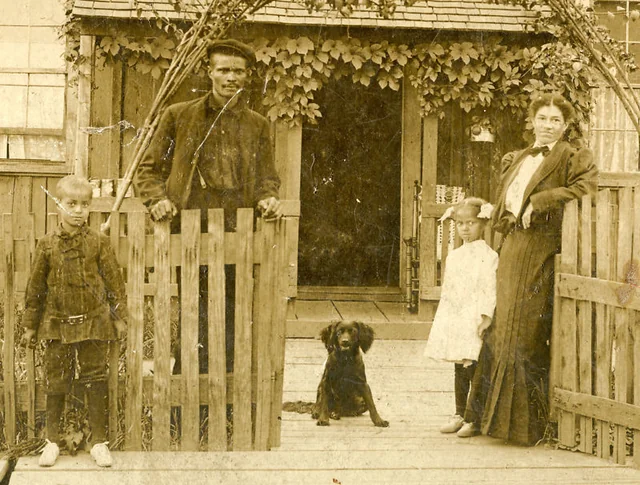 An old sepia photo shows a Black family of four and a dog posing outside a wooden house. The father and son stand by an open gate, while the mother and daughter stand on the porch. Ivy climbs up the house behind them.