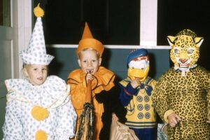 Four children in colorful Halloween costumes stand indoors. From left to right: a clown, a pumpkin, a character with a Donald Duck mask, and a child in a leopard costume with a full face mask.