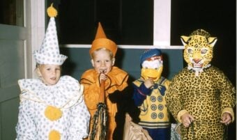 Four children in colorful Halloween costumes stand indoors. From left to right: a clown, a pumpkin, a character with a Donald Duck mask, and a child in a leopard costume with a full face mask.