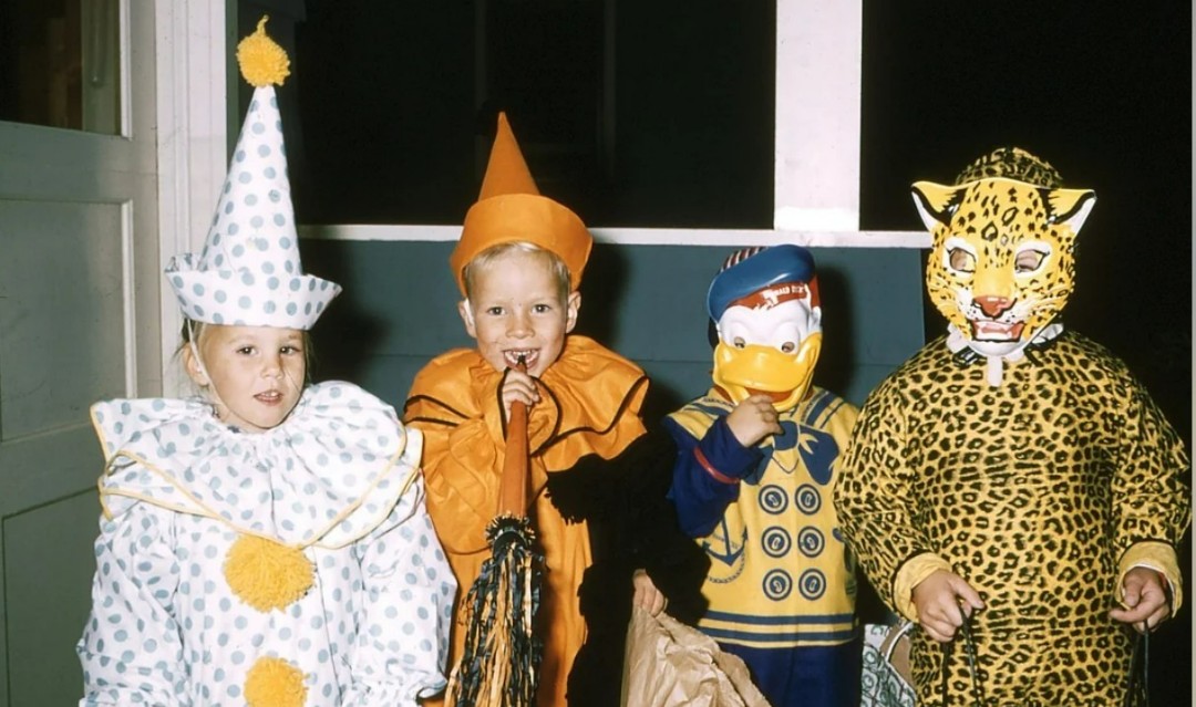 Four children in colorful Halloween costumes stand indoors. From left to right: a clown, a pumpkin, a character with a Donald Duck mask, and a child in a leopard costume with a full face mask.