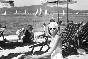 A woman in a swimsuit and white sunglasses sits on a beach chair under an umbrella. Several people relax on the sandy beach and sailboats are visible on the water, with mountains in the distance. The image is black and white.