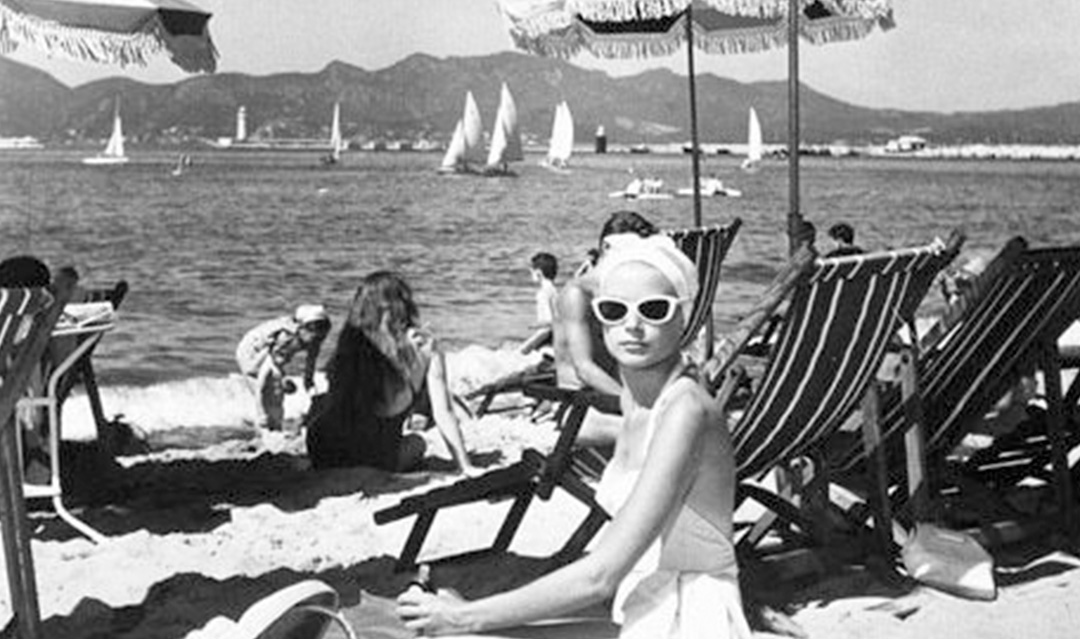 A woman in a swimsuit and white sunglasses sits on a beach chair under an umbrella. Several people relax on the sandy beach and sailboats are visible on the water, with mountains in the distance. The image is black and white.