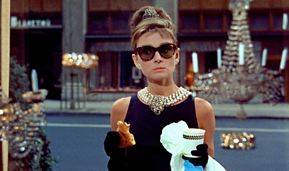 A woman with a beehive updo, tiara, pearl necklace, black dress, and sunglasses stands on a city street holding a pastry and a cup, with gloved hands and a shop window in the background.