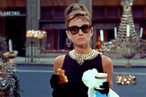 A woman with a beehive updo, tiara, pearl necklace, black dress, and sunglasses stands on a city street holding a pastry and a cup, with gloved hands and a shop window in the background.