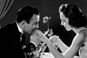 A man in a suit kisses a woman's hand across a candlelit dinner table. The woman, in a sleeveless dress, smiles softly. A rose in a vase and a curtain are visible in the background. The image is in black and white.