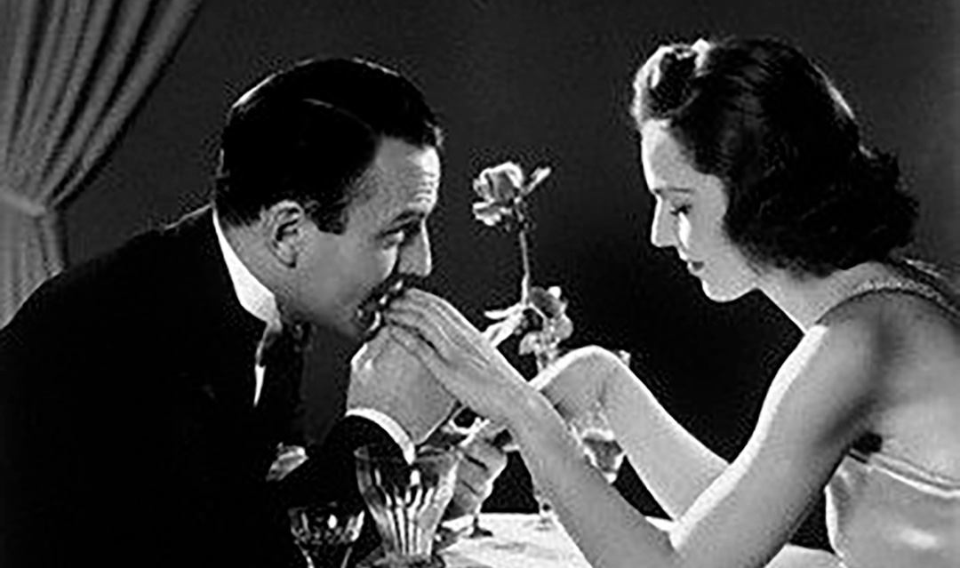 A man in a suit kisses a woman's hand across a candlelit dinner table. The woman, in a sleeveless dress, smiles softly. A rose in a vase and a curtain are visible in the background. The image is in black and white.