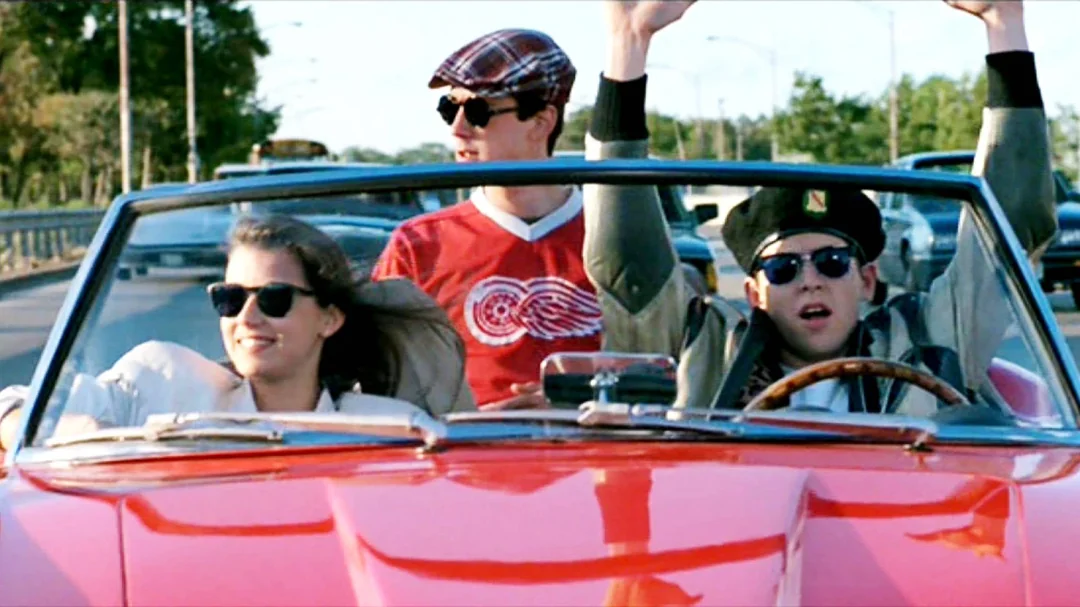 Three young people wearing sunglasses ride in a red convertible with the top down. One person drives, another smiles in the passenger seat, and the third stands up in the back, arms raised, enjoying the ride on a sunny day.