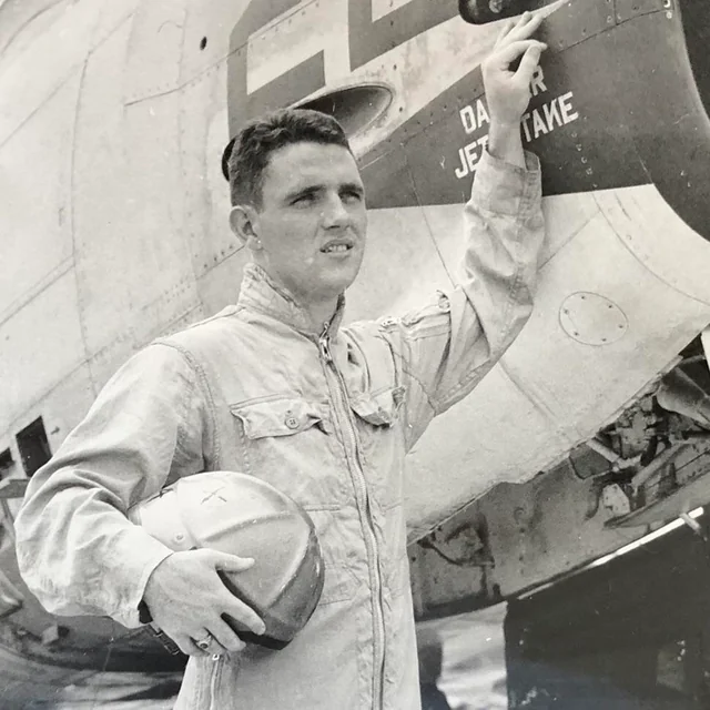 A man in a flight suit holds a helmet and stands beside an aircraft, pointing to a warning label that reads “DANGER JET INTAKE.” The plane’s metal body is visible in the background. The image is black and white.