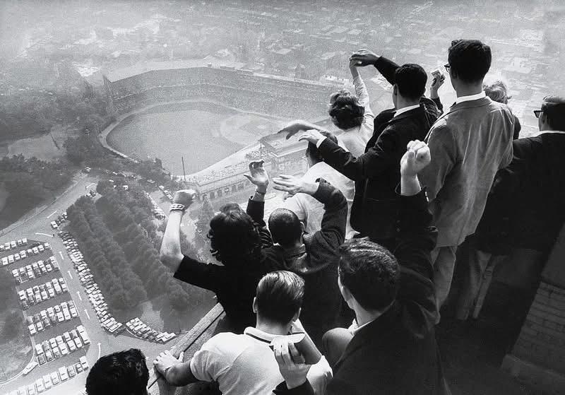 A group of people on a rooftop excitedly wave and cheer while overlooking a crowded baseball stadium below, with cars parked outside and a cityscape visible in the distance.