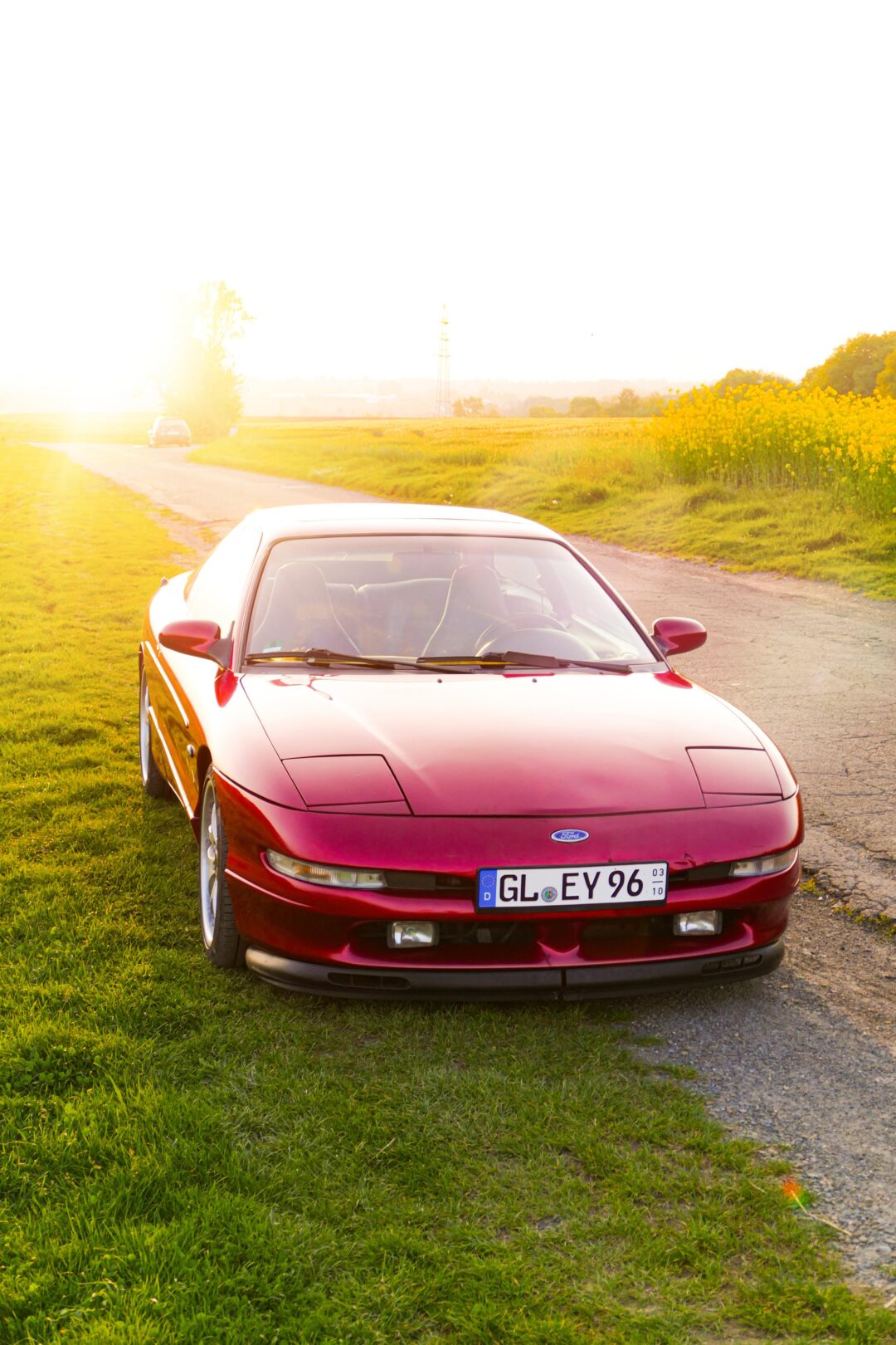 A red Ford Probe parked on grassy roadside at sunset, with sunlight reflecting off its hood and a rural landscape in the background.