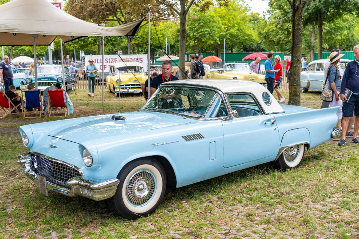 A light blue vintage car with a white roof is parked on grass at an outdoor car show. People stand nearby, and other classic cars are visible in the background under trees.
