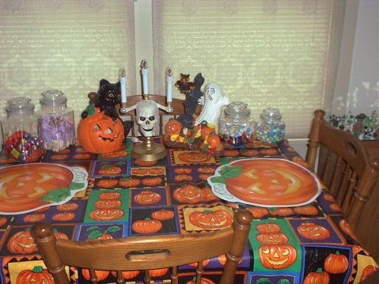 A dining table decorated for Halloween with a pumpkin-patterned tablecloth, matching pumpkin placemats, Halloween figurines, jars of candy, and two wooden chairs in front of a window with closed blinds.