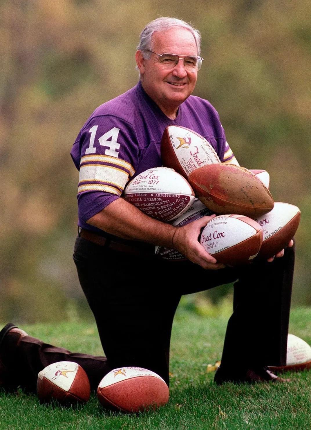 An older man wearing a purple football jersey with number 14 kneels on grass, smiling and holding several footballs in his arms, with more footballs on the ground beside him.