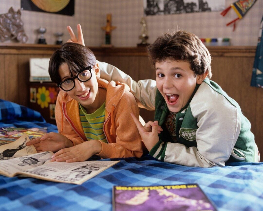 Two boys are lying on a bed in a cozy, colorful room. One boy in glasses reads a comic while the other, smiling widely, gives him bunny ears. Comics are scattered on the bed, and trophies are visible in the background.