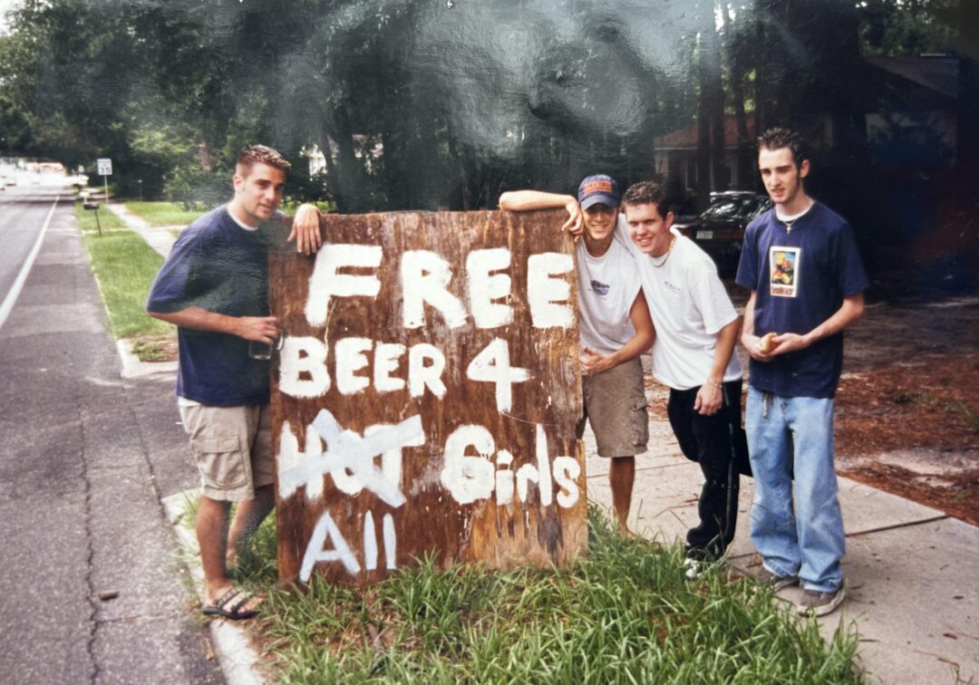 Four young men stand on a sidewalk next to a large, homemade wooden sign that reads "FREE BEER 4 HOT Girls All" in white paint. Trees, grass, and a road are visible in the background.