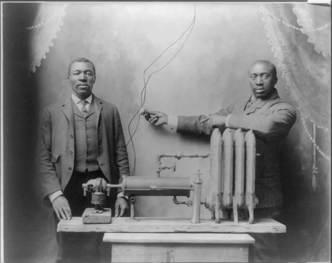 Two Black men in suits stand beside a mechanical device on a wooden table, one man holding wires attached to it. The background is a studio setting with decorative curtains.