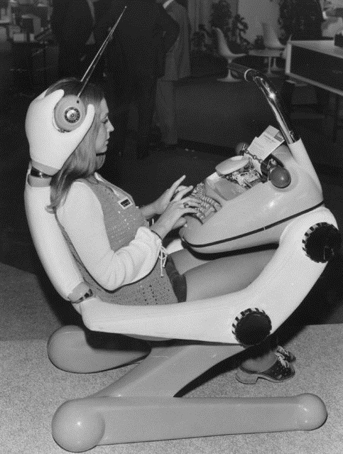 A woman sits in a futuristic, pod-shaped chair that resembles a machine, typing on an integrated typewriter. The chair has a visor, control panel, and large mechanical joints. The scene appears to be from a past technology exhibition.