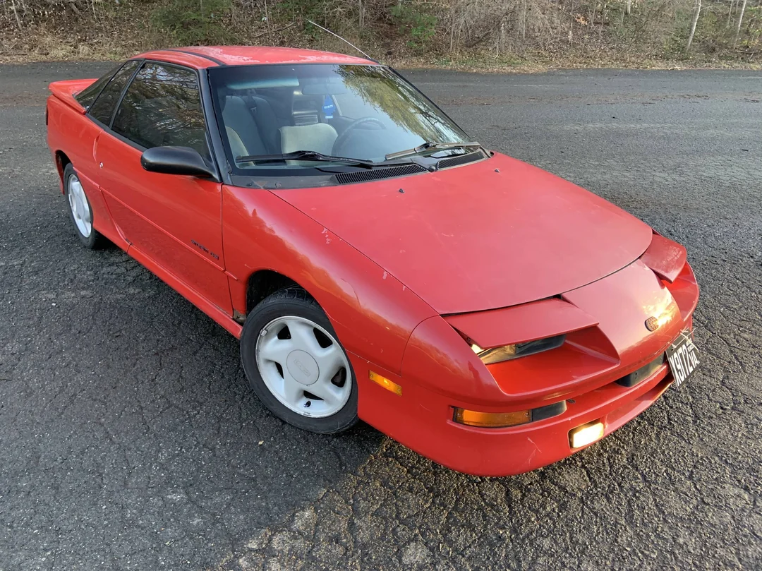 A red 1990s sports car with pop-up headlights, white wheels, and a slightly faded hood is parked on a cracked asphalt road near some dry grass and trees.