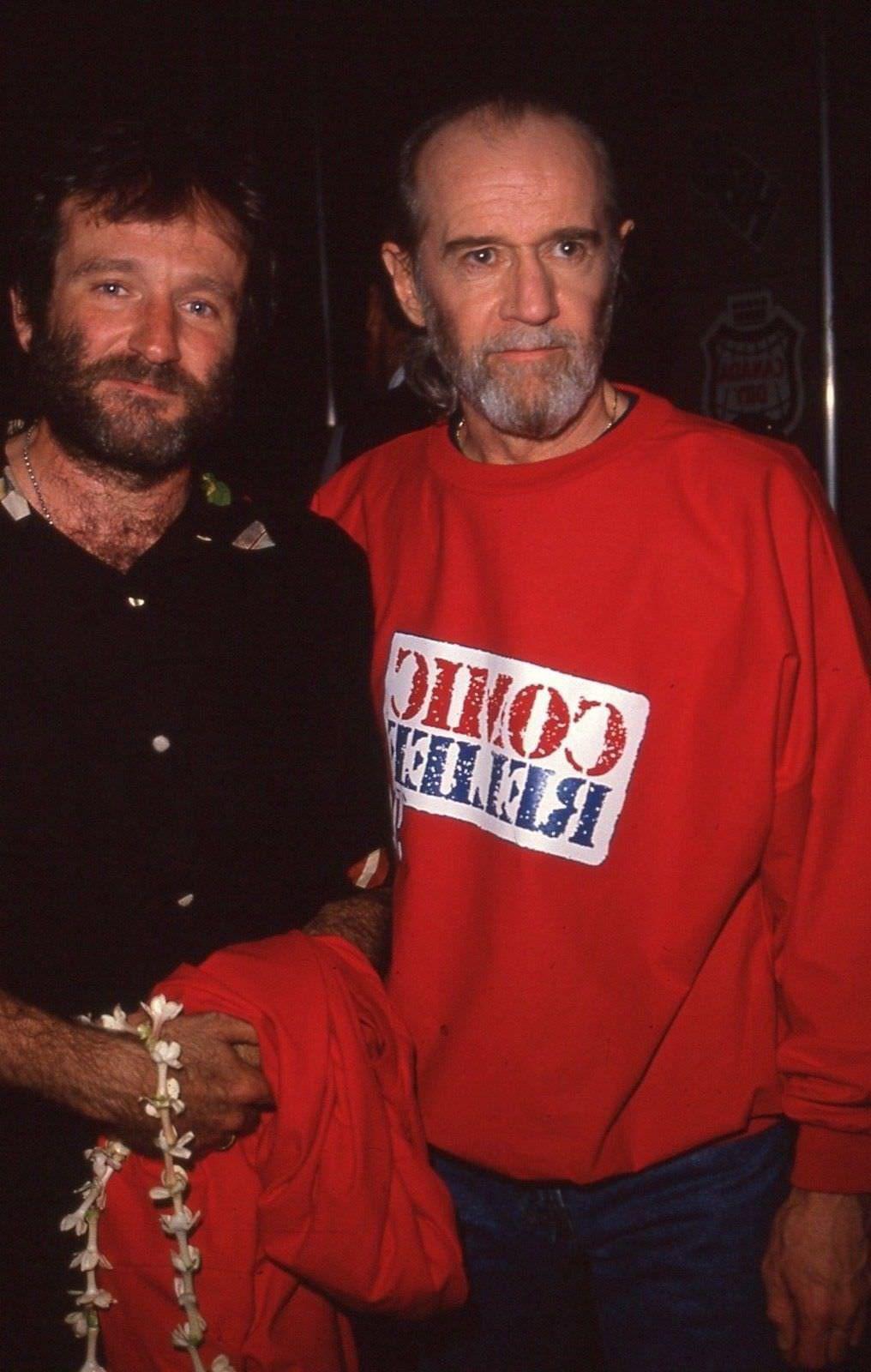 Two men stand together. One wears a black shirt and holds a red jacket and a flower lei, while the other wears a red sweatshirt with text printed in white and blue. Both have beards and look at the camera in a dimly lit setting.