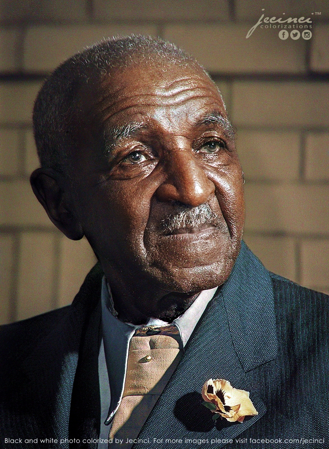 Elderly man with gray hair and a mustache, wearing a dark suit with a tan shirt and gold flower pin, looking thoughtfully into the distance against a blurred, neutral background.