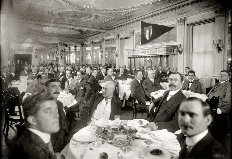 A large group of men in suits sit at round tables set for a formal meal in an elegant, ornate dining room. A flag with a shield emblem hangs on the wall, and handwritten text is visible at the top of the photo.