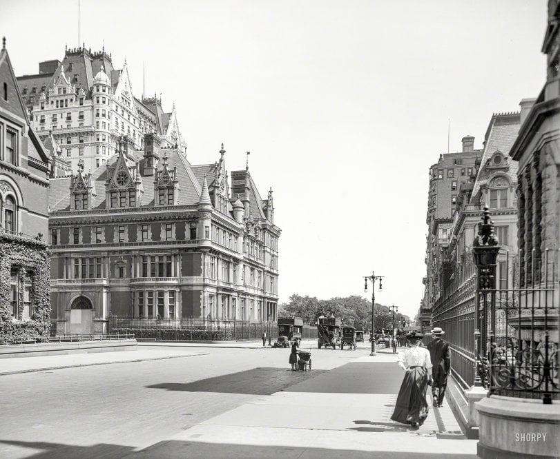 A black and white photo shows a wide city street lined with ornate buildings, horse-drawn carriages, and people in early 20th-century clothing walking on the sidewalk.