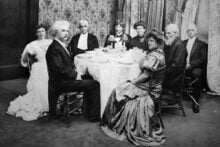 Eight people in formal attire sit around a round dining table in an ornate room with patterned wallpaper and curtains, looking toward the camera. The table is set for a formal meal.