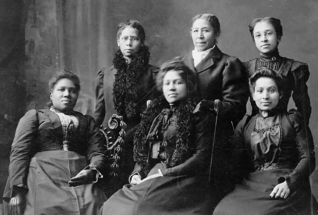 Six Black women in formal late 19th-century clothing pose together for a studio portrait, with three seated in front and three standing behind, all looking seriously at the camera.