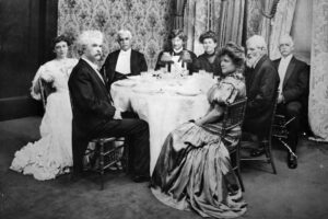 Eight people in formal attire sit around a round dining table in an ornate room with patterned wallpaper and curtains, looking toward the camera. The table is set for a formal meal.