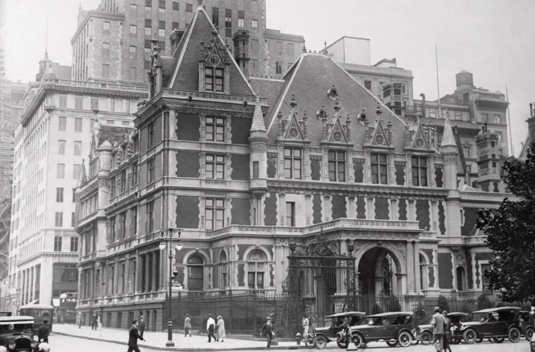 Historic black-and-white photo of a grand, ornate mansion with steep roofs and pointed gables, surrounded by early 20th-century cars and people walking along the street. Tall city buildings rise in the background.