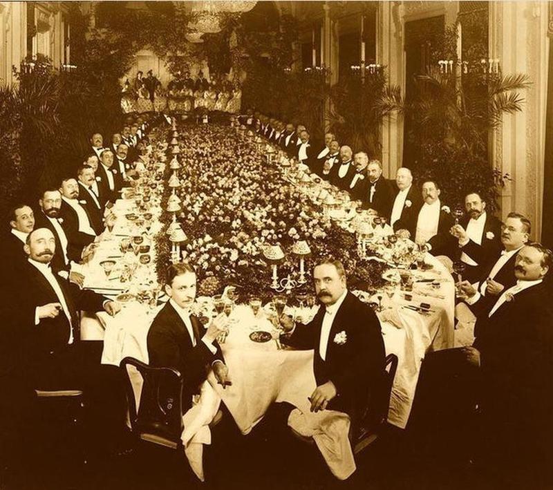 A sepia-toned, historical photo shows many men in formal attire seated around a long, ornate banquet table, adorned with flowers and glassware, in an opulent hall with chandeliers and tall plants.