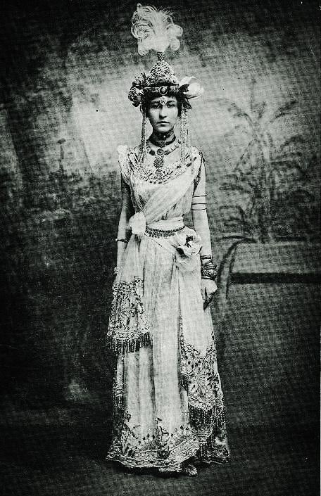 A woman stands facing forward, wearing an ornate, traditional costume with intricate patterns, jewelry, and a detailed headdress with feathers. The image is black and white with a vintage, historical atmosphere.