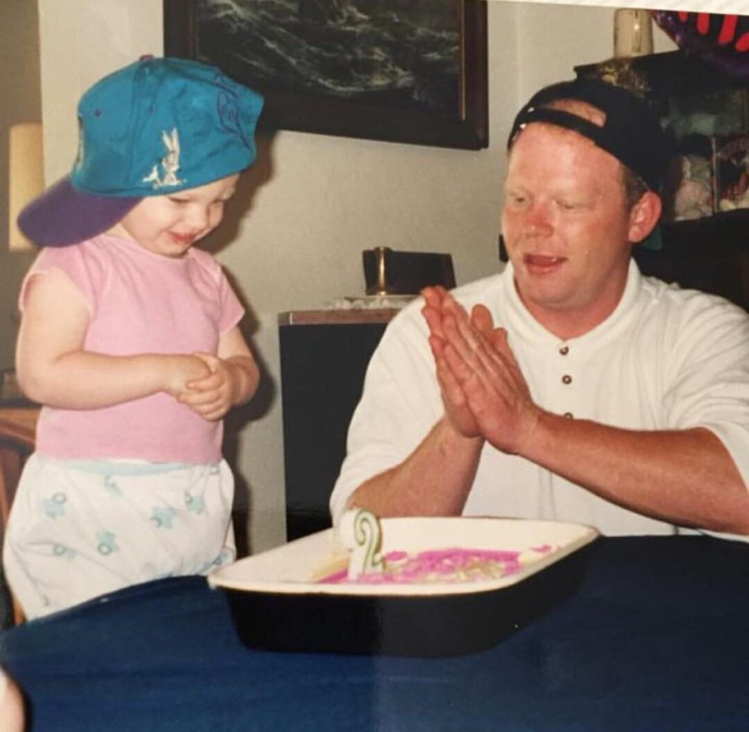 A smiling child in a blue sideways cap and an adult in a backwards black cap sit at a table with a rectangular birthday cake, which has a green number 2 candle on it. The adult is clapping while the child looks on happily.