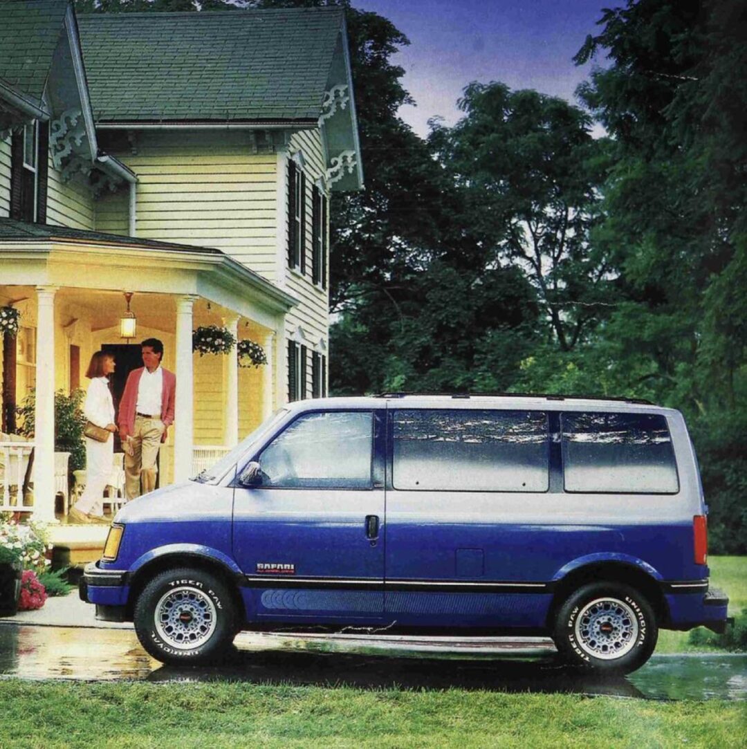 A blue and white minivan is parked on a driveway in front of a yellow house with a porch; two people stand on the porch, talking, with green trees in the background.