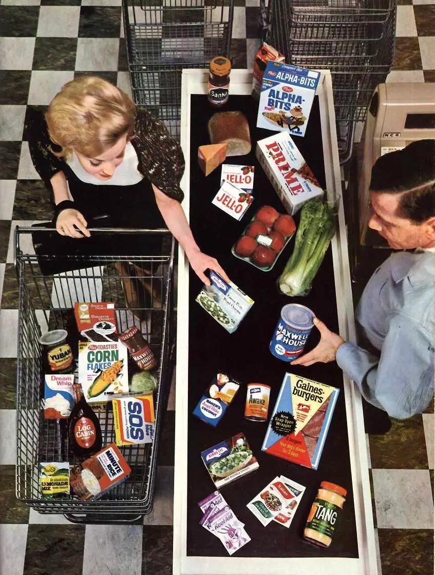 A woman loads groceries onto a checkout conveyor belt while a cashier scans items. The cart and belt are filled with various 1960s food products in colorful packaging. The scene is in a grocery store with checkered tile flooring.