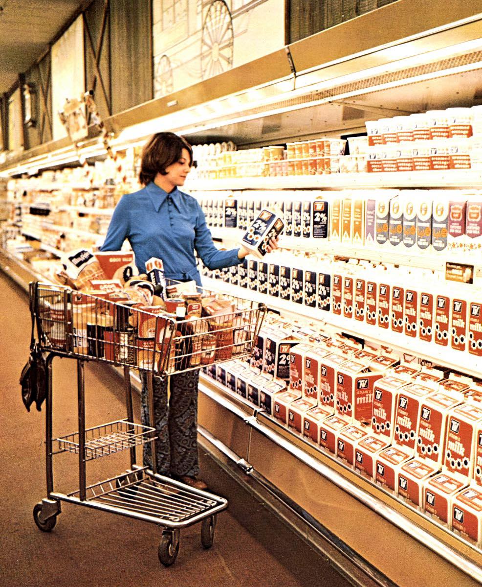 A woman in a blue shirt stands in a grocery store aisle, holding a carton of milk and shopping with a cart filled with groceries. Shelves are lined with various milk and dairy products.