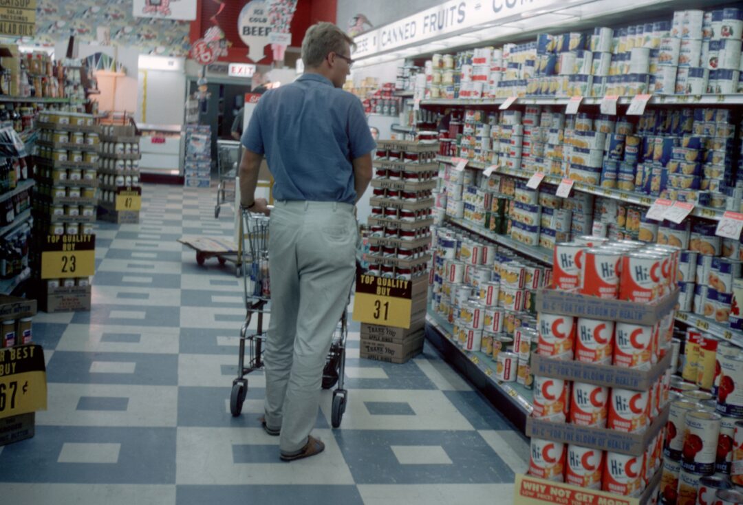 A man in a blue shirt and light pants pushes a shopping cart down a grocery store aisle lined with shelves full of canned goods and colorful labels. The store has checkered floors and overhead fluorescent lights.