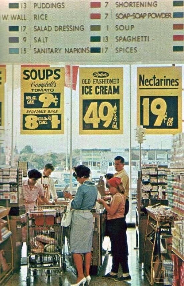Shoppers with carts check out at a grocery store in front of large yellow sale signs for soup, ice cream, and nectarines. Above, a board lists food items and aisle numbers. Store windows show a parking lot outside.