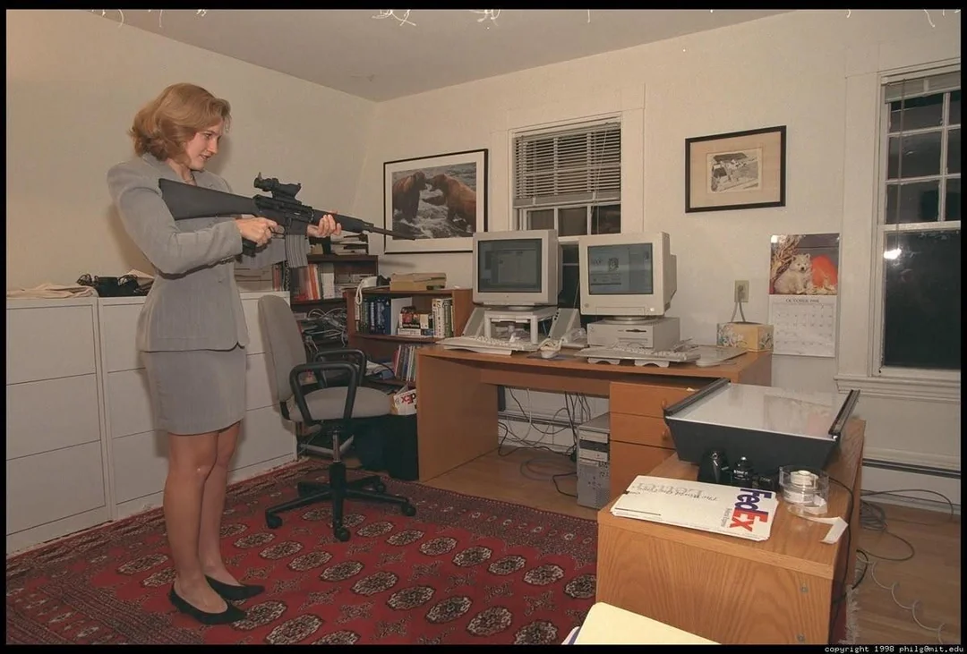 A woman in a gray suit stands in an office aiming a large rifle. The room has desks with two computer monitors, office chairs, bookshelves, a calendar, and framed pictures on the wall.