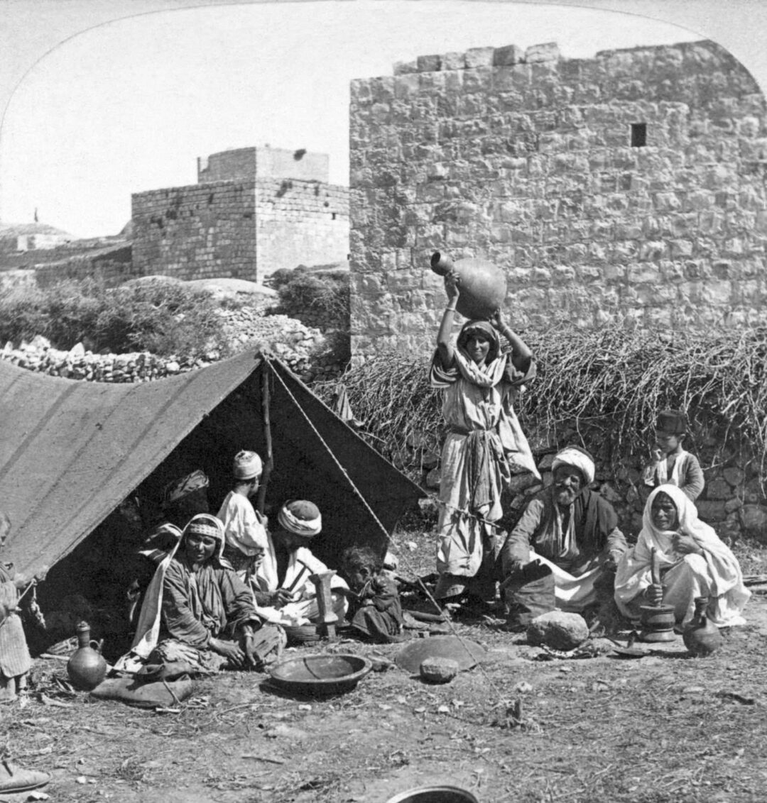 A group of people in traditional clothing sit and stand outside a tent near stone buildings. One woman balances a jug on her head while others sit on the ground with pottery and household items scattered nearby.