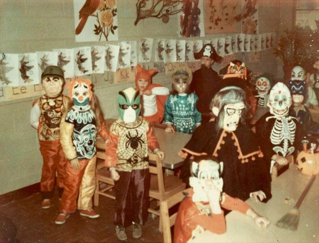 A group of children in colorful Halloween costumes and masks sit and stand around tables in a classroom decorated with artwork and drawings on the walls.