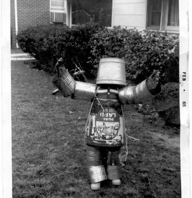 A child in a homemade robot costume made from metal cans and buckets stands outside on grass with arms raised. The photo appears old, dated February 1965, with houses and bushes in the background.
