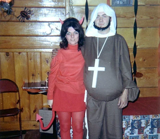 A woman dressed as a devil stands next to a man dressed as a monk with a large cross necklace. They are posing indoors at a party decorated with streamers, wood-paneled walls, and a folding chair.
